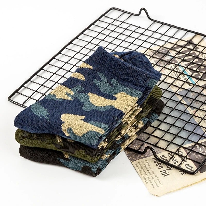 A pair of comfortable Camo socks by BlasterMaster in various shades are neatly folded and placed on a black wire mesh alongside a partially visible newspaper.