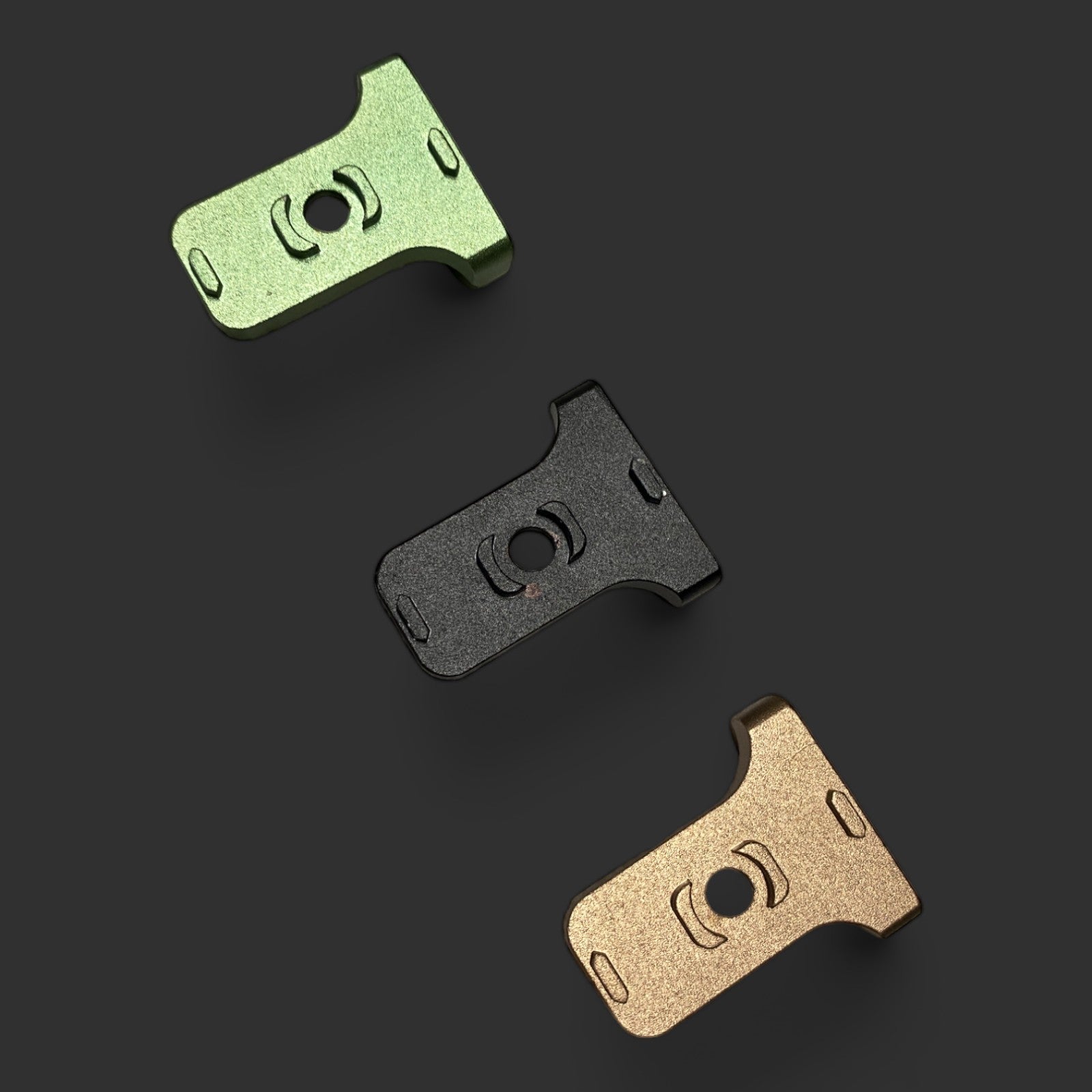 Three MLOCK CQD MLOK Forward Sling Mounts in green, black, and bronze are displayed on a dark surface. The mounts boast a unique cutout design, perfect for tactical performance, and appear identical in shape and size despite their differing colors.