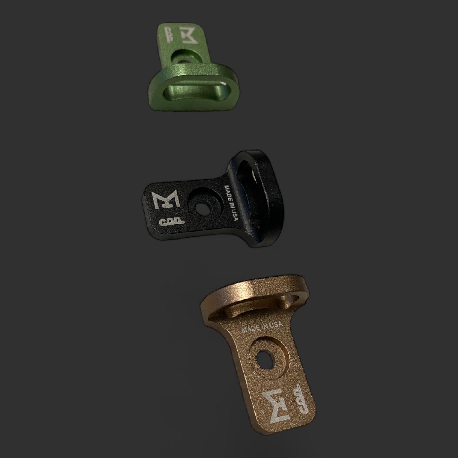 Three metal loop attachment parts in green, black, and bronze are arranged in a vertical line on a dark background. Each piece has inscriptions and logos indicating they are made in the USA by MLOCK, showcasing tactical performance and CQD MLOK Forward Sling Mount compatibility.